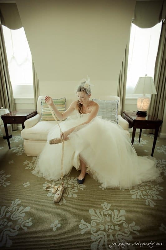 carolina inn wedding photography ~ danielle and thomas A bride in a strapless white gown and birdcage veil sits on a sofa in a room with light-colored decor. She holds a pair of ballet shoes by the ribbons, looking at them thoughtfully.