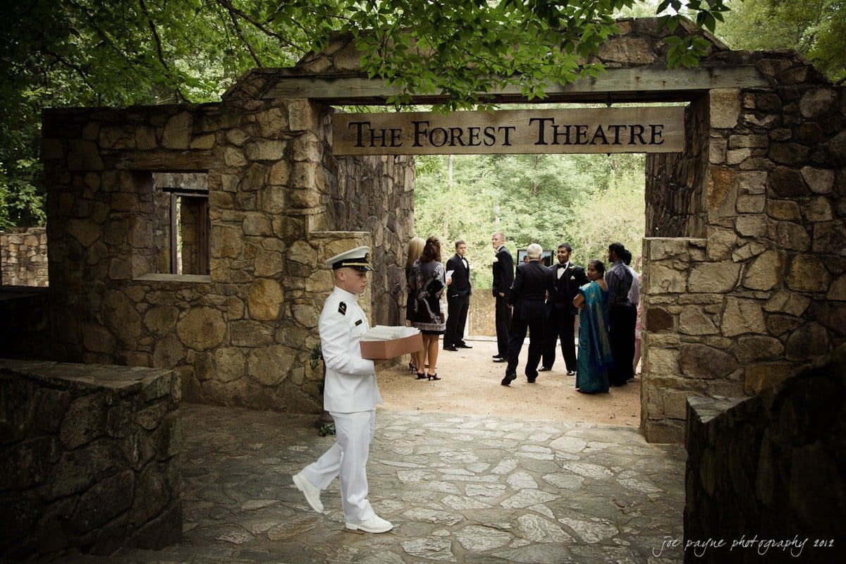 carolina inn wedding photography ~ danielle and thomas A person in a naval uniform carries a box past a stone archway marked The Forest Theatre. In the background, a group of people gather, some wearing formal attire. The scene is set outdoors among trees and stone structures.