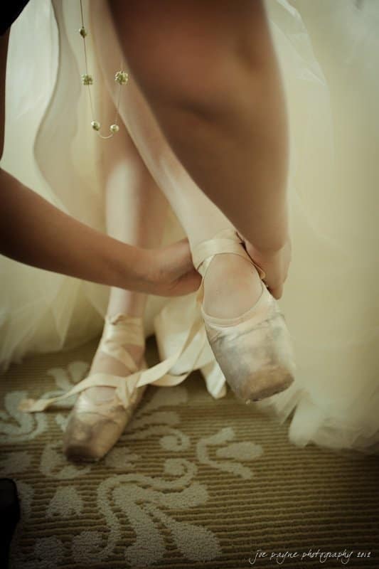 carolina inn wedding photography ~ danielle and thomas A dancer in a white dress is having her ballet shoes adjusted by another person, preparing for a performance. The focus is on the pointe shoes and the hands tying the ribbons. The scene is softly lit, creating an elegant atmosphere.