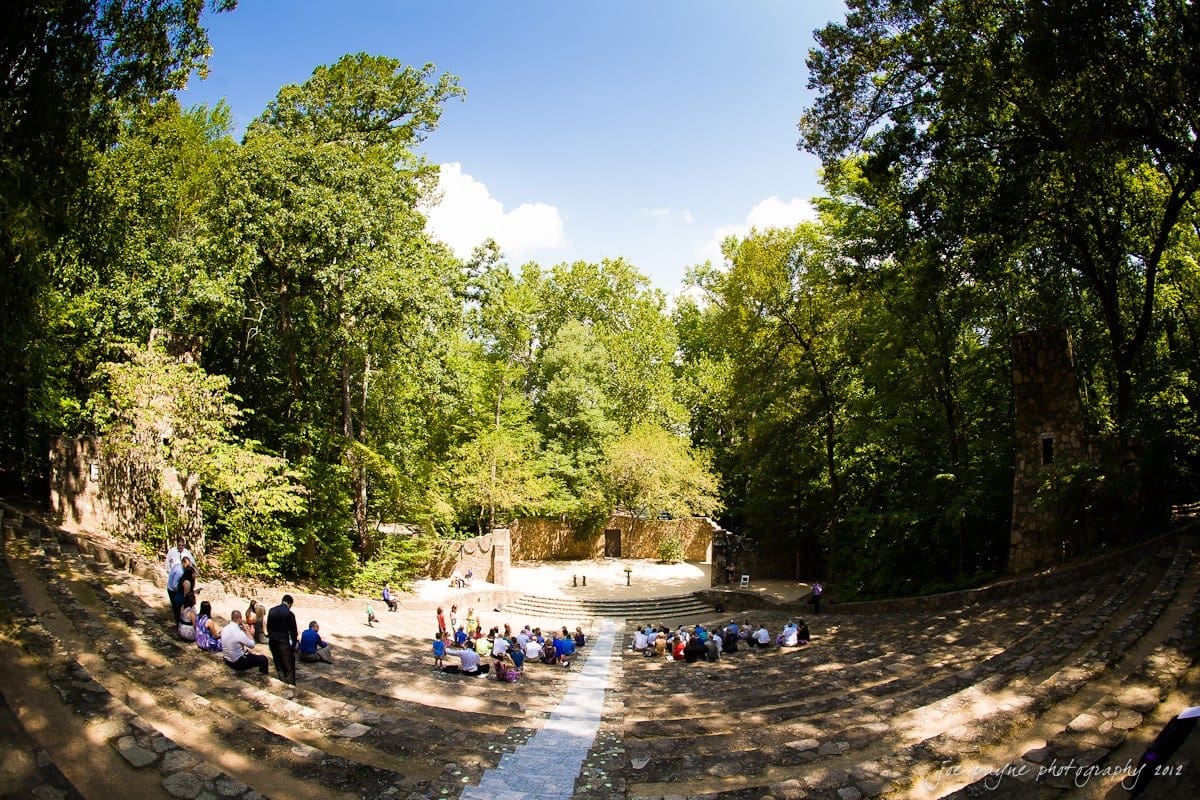 carolina inn wedding photography ~ danielle and thomas Wide-angle view of an outdoor amphitheater surrounded by lush trees. A few people are seated on stone steps, watching the central stage area. The scene is under a clear blue sky with fluffy clouds.