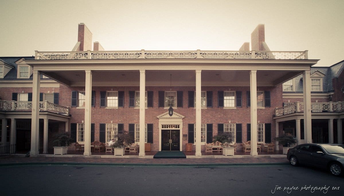 carolina inn wedding photography ~ danielle and thomas A large brick building with white pillars and a porch spans the front. The windows have dark shutters, and chairs are arranged on the porch. The sky is clear. A small sign above the entrance bears the name of the establishment.