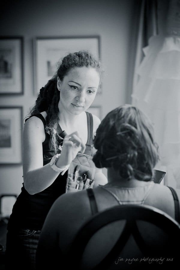 A makeup artist with long hair and a brush in hand is applying makeup to a seated woman in a dark top. The scene is in a softly lit room with framed pictures on the wall in the background.