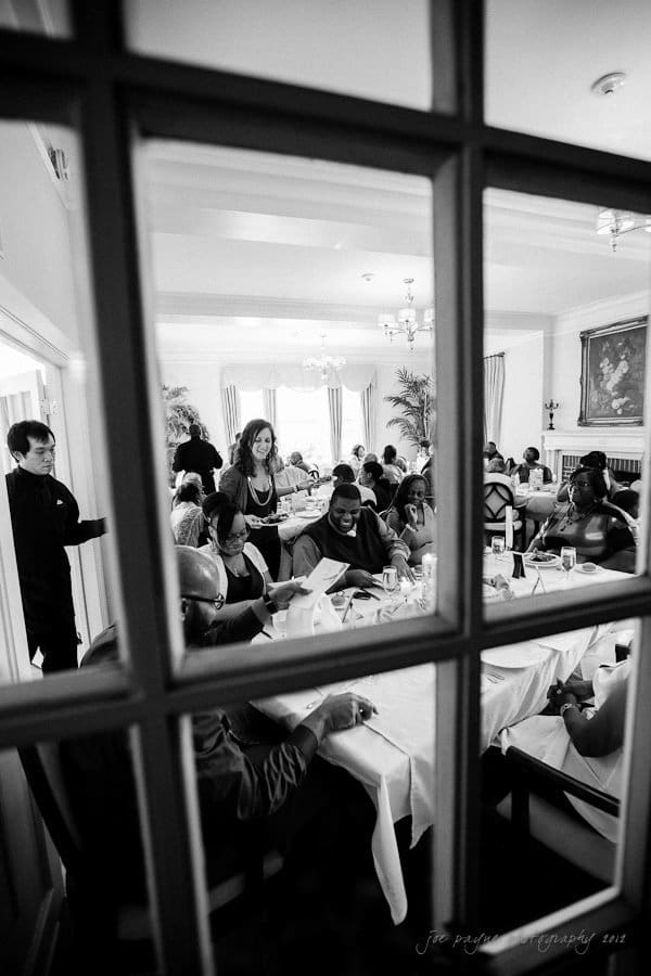 A black and white photo of people gathered in a dining room, seated at tables with white tablecloths. A waitress stands near the center. The scene is viewed through a window pane.