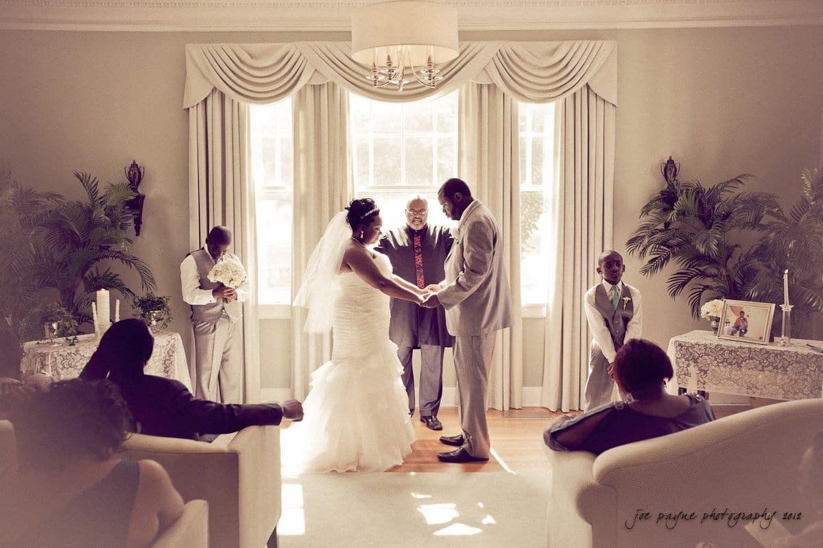 A couple holds hands during their wedding ceremony in a bright room. The officiant stands between them, while two young attendants are nearby. Guests are seated on white chairs, and sunlight streams in through a large window with curtains.