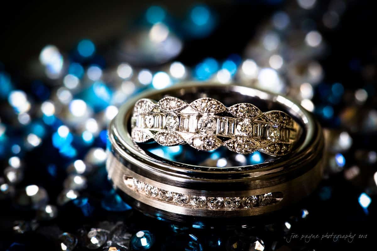 Close-up of two elegant rings on a sparkling blue and black surface. The top ring features intricate detailing with small diamonds, while the bottom ring has a sleek design with a row of diamonds embedded around it.