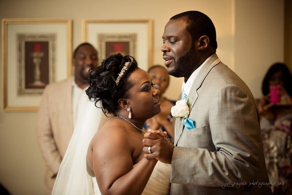 A couple is joyfully dancing at their wedding, surrounded by smiling guests. The bride wears a white gown with a veil, and the groom is in a light gray suit with a blue tie and a white rose boutonniere. Framed art decorates the wall behind them.