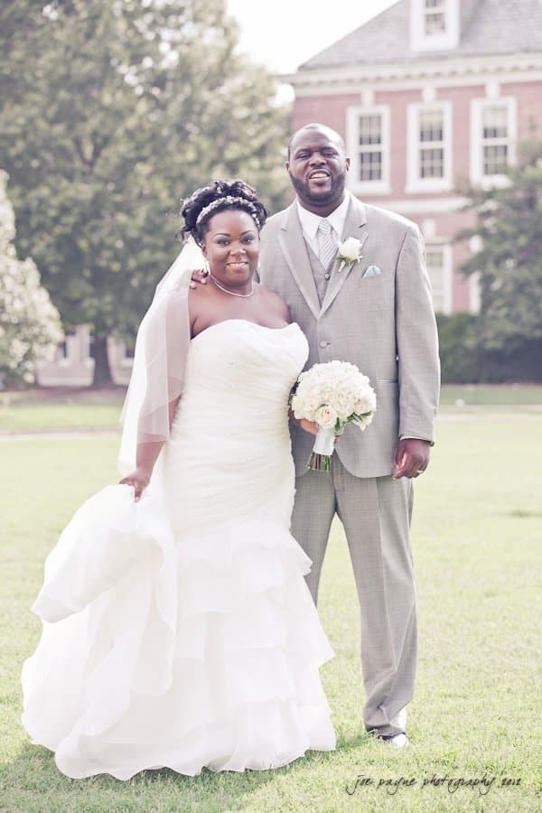 A bride and groom smile while standing on a grassy area in front of a brick building. The bride wears a strapless white gown and holds a bouquet, the groom wears a light gray suit. Both are looking at the camera.