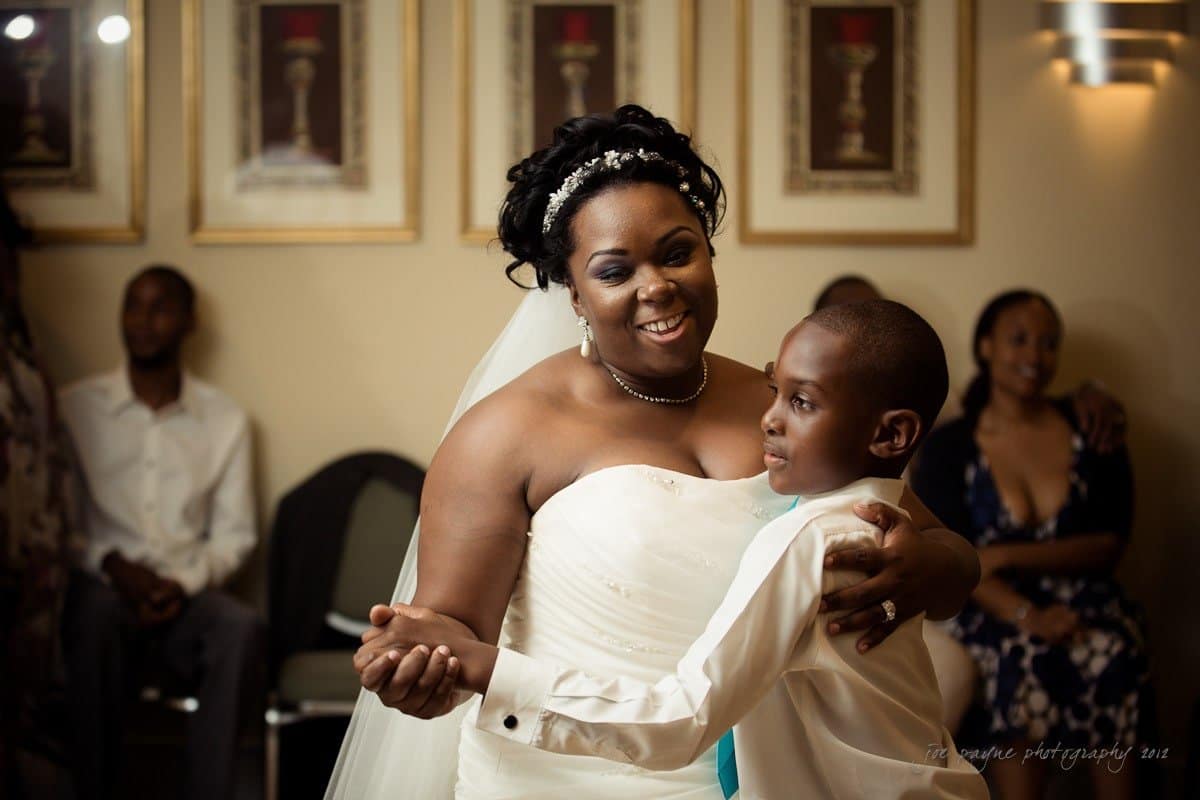 A bride in a white dress and veil dances joyfully with a young boy in a shirt and tie. They are smiling in a warmly lit room with framed pictures on the wall. Other guests are seated and watching in the background.