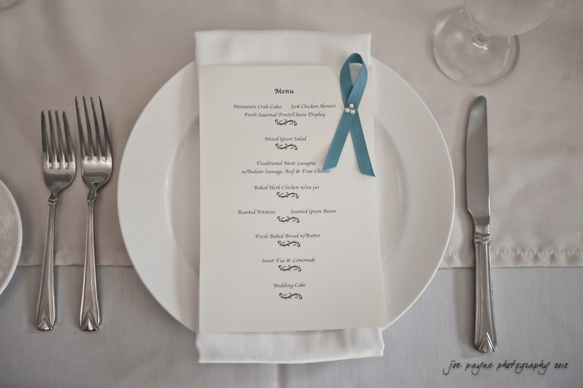 A neatly arranged place setting with a menu on a white plate. The menu has a blue ribbon and includes a list of items like salmon, chicken, pasta, cakes, and drinks. Silver cutlery is placed on either side of the plate. The tablecloth is light gray.