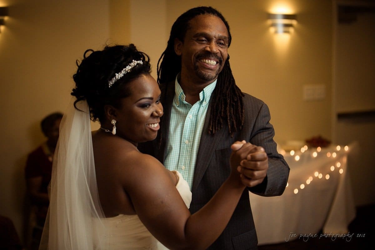 A bride and a man with long hair are joyfully dancing indoors. The bride is wearing a strapless white dress and veil, while the man is in a suit. The background features warm lighting and a softly lit room.