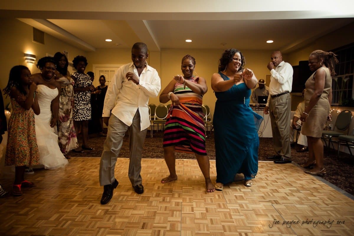 A group of people dancing joyfully on a wooden dance floor at a party or celebration. They are dressed in formal and semi-formal attire, and there is a festive, cheerful atmosphere in the room.