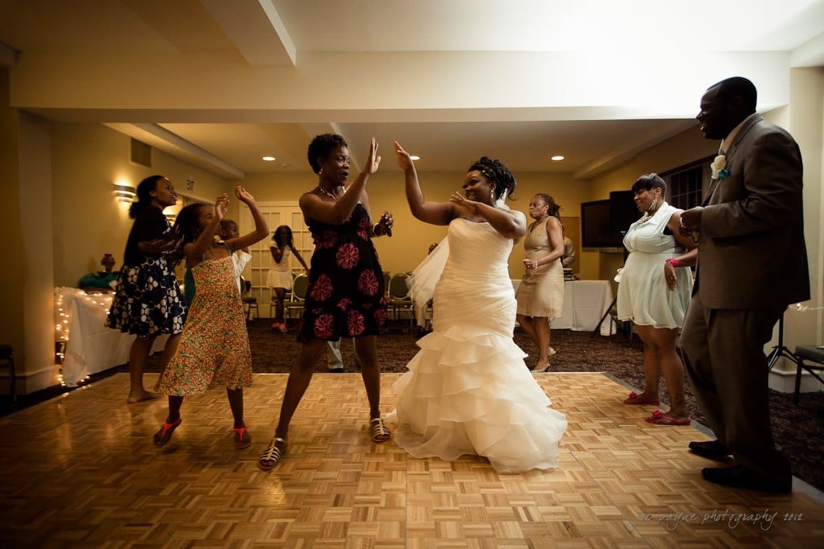 A bride in a white wedding dress dances joyfully on a wooden floor with a group of people. Everyone is celebrating, dressed in colorful attire. The atmosphere is lively and festive, with some guests clapping and others dancing along.