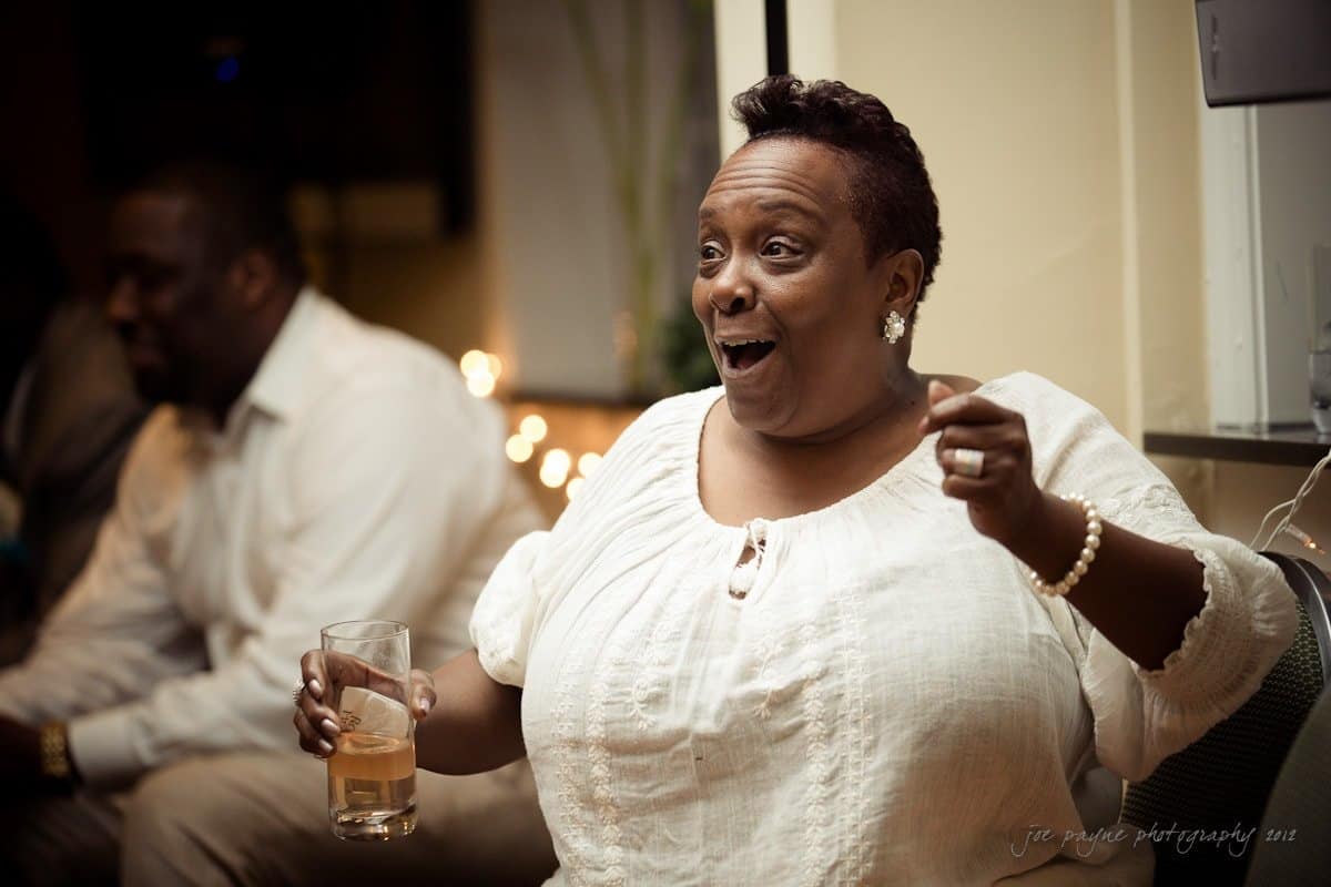 A woman in a white blouse is smiling and holding a drink, looking surprised or delighted, with a blurred person in the background. The setting appears to be indoors with warm lighting.