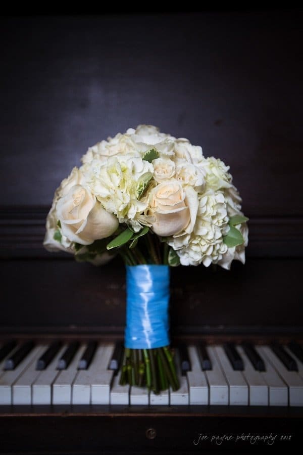 A bouquet of white and cream roses, wrapped with blue ribbon, is placed on the keys of a dark piano.