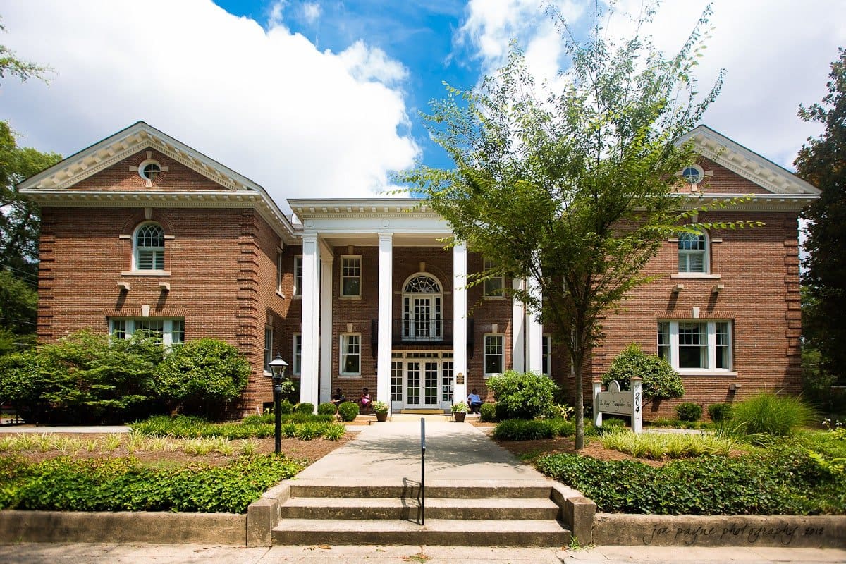 A symmetrical two-story brick building with a central entrance flanked by tall white columns. It features arched windows and is surrounded by greenery and trees, set against a partly cloudy blue sky.
