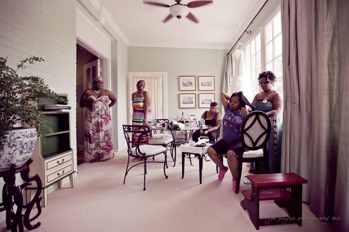 A group of women enjoying a relaxed moment in a living room. Some are seated, and others are standing, engaging in conversation. The room has chairs, a table with items on it, and a large window with curtains, allowing natural light to enter.