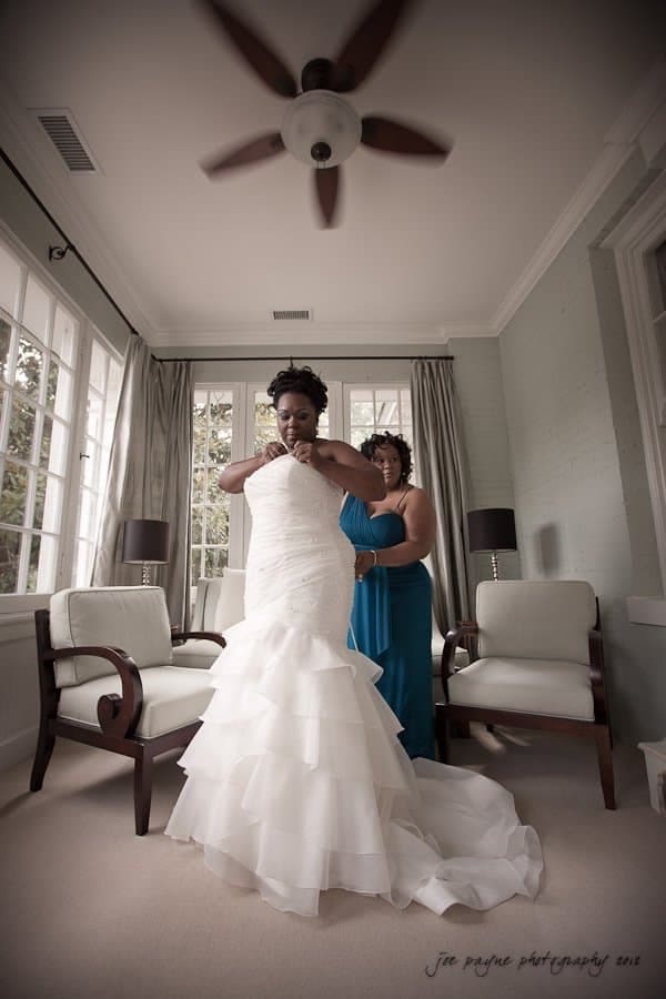 A bride in a white gown stands in a room with large windows, being assisted by a woman in a blue dress. The room is elegantly decorated with chairs and curtains, and a ceiling fan is visible above.