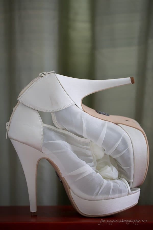 A pair of elegant white high heels with a sheer, gathered fabric detail on the sides, displayed on a wooden surface against a muted green background.