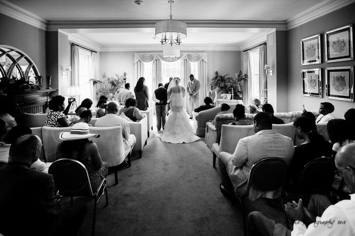 Black and white photo of an indoor wedding ceremony. The bride and groom stand at the front of a room filled with guests seated on chairs and sofas. Large windows and curtains frame the room, and plants decorate the space.