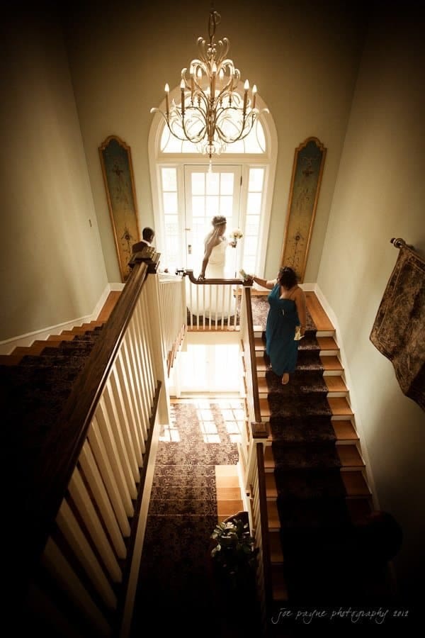 A bride in a white dress descends a staircase with two other people, one in a blue dress, captured in a warmly lit, elegant interior with a chandelier and large window.