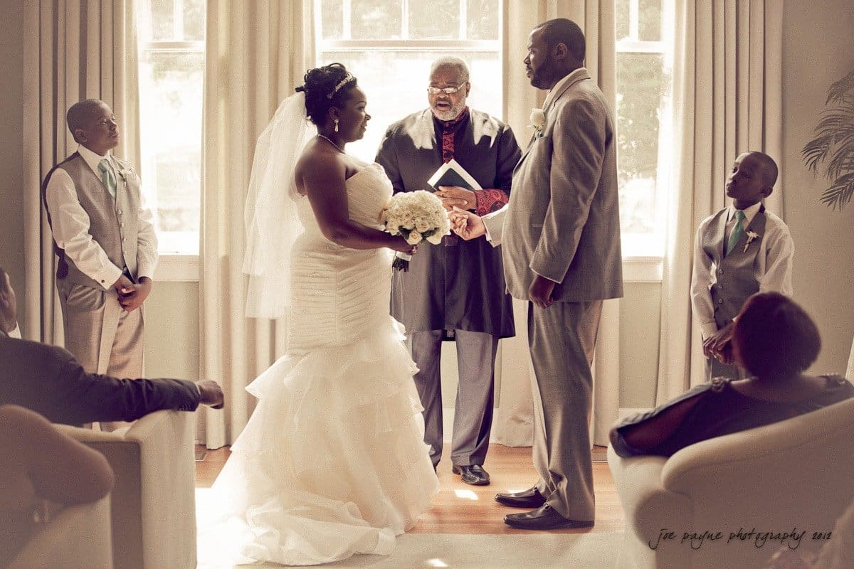 A bride and groom stand facing each other during a wedding ceremony, with a man officiating. Two young boys in suits stand nearby. Many of the happy attendees watch from their seats in a warmly lit room with large windows.