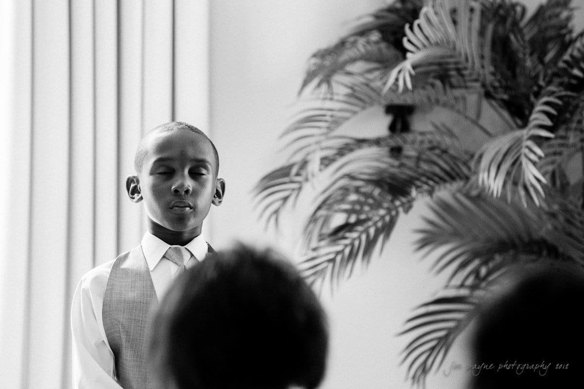 Black and white image of a young boy standing with eyes closed, wearing a vest and tie. He appears to be speaking or singing. Behind him is a large potted plant with long leaves.