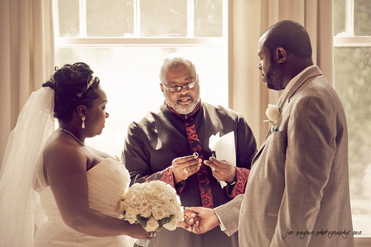 A couple stands before an officiant during a wedding ceremony. The bride holds a bouquet of white roses and wears a white gown with a veil. The groom is in a light-colored suit with a boutonniere. The officiant reads from a book, solemnly conducting the ceremony.