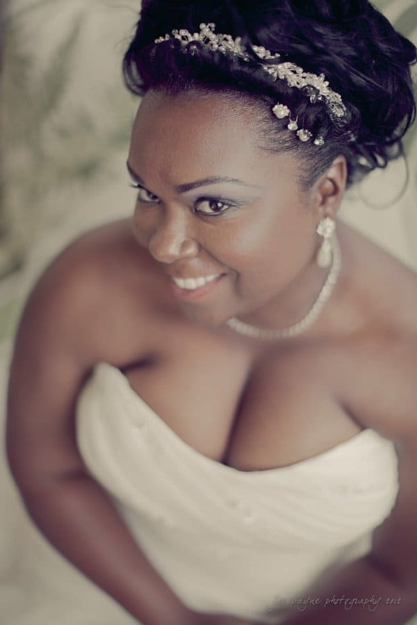 A bride in a strapless white dress smiles at the camera. She wears a jeweled tiara in her hair and matching earrings. The photo is taken from a slightly elevated angle, focusing on her joyful expression.