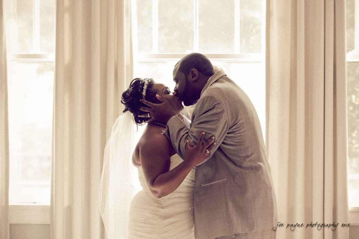 A newlywed couple kisses in front of bright windows with beige curtains. The bride wears a strapless white gown and veil, and the groom wears a light gray suit. They hold each others faces lovingly.
