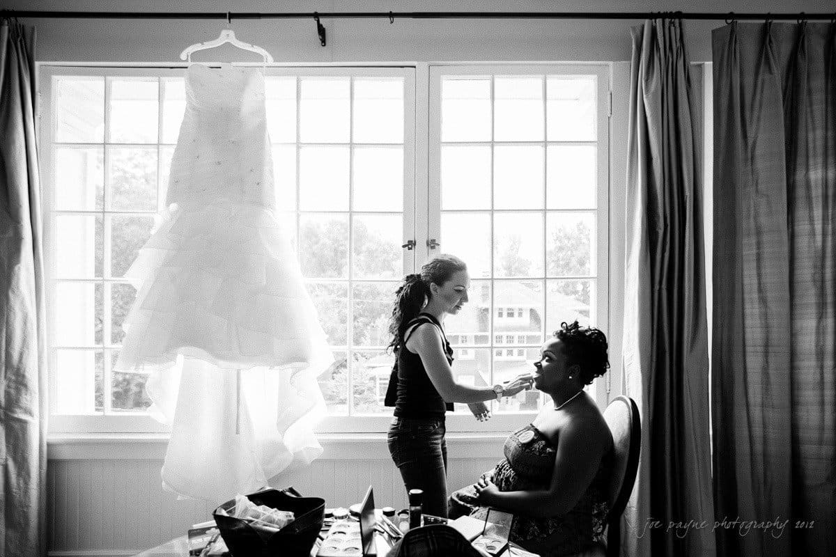 A woman seated in a room gets her makeup done by another woman standing next to her. A wedding dress hangs in front of a large window that fills the room with light. A table nearby holds various items.