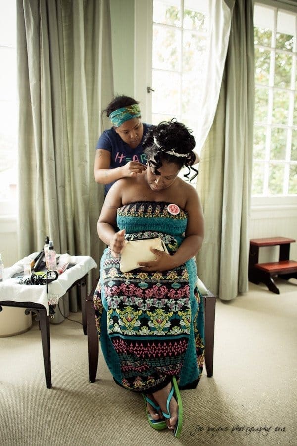 A woman sits on a chair in a brightly lit room with large windows, while a young girl styles her hair. The woman wears a colorful patterned dress and holds a small pouch. A table with hair styling tools is nearby.