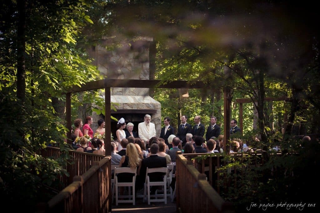 A wedding ceremony outdoors in a serene, wooded area. Guests are seated on white chairs facing a couple standing in front of a rustic stone fireplace. Trees surround the venue, creating a natural, intimate atmosphere.