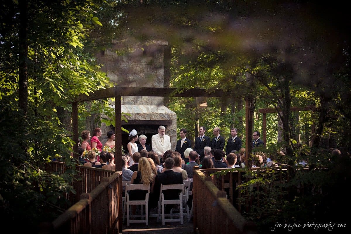 barn at valhalla, chapel hill wedding photography ~ allegra and chris A wedding ceremony outdoors in a serene, wooded area. Guests are seated on white chairs facing a couple standing in front of a rustic stone fireplace. Trees surround the venue, creating a natural, intimate atmosphere.