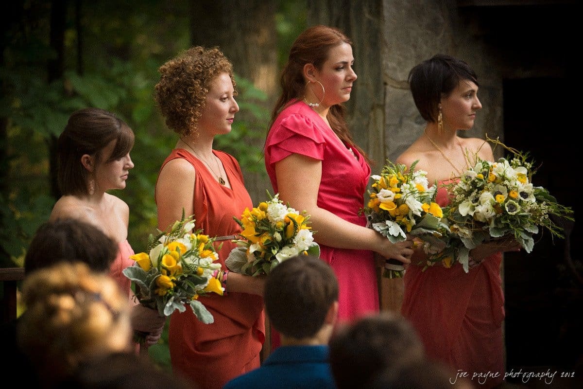 barn at valhalla, chapel hill wedding photography ~ allegra and chris Four women in colorful dresses stand side by side holding vibrant floral bouquets. They are outside, with a blurred natural background of trees and stone.