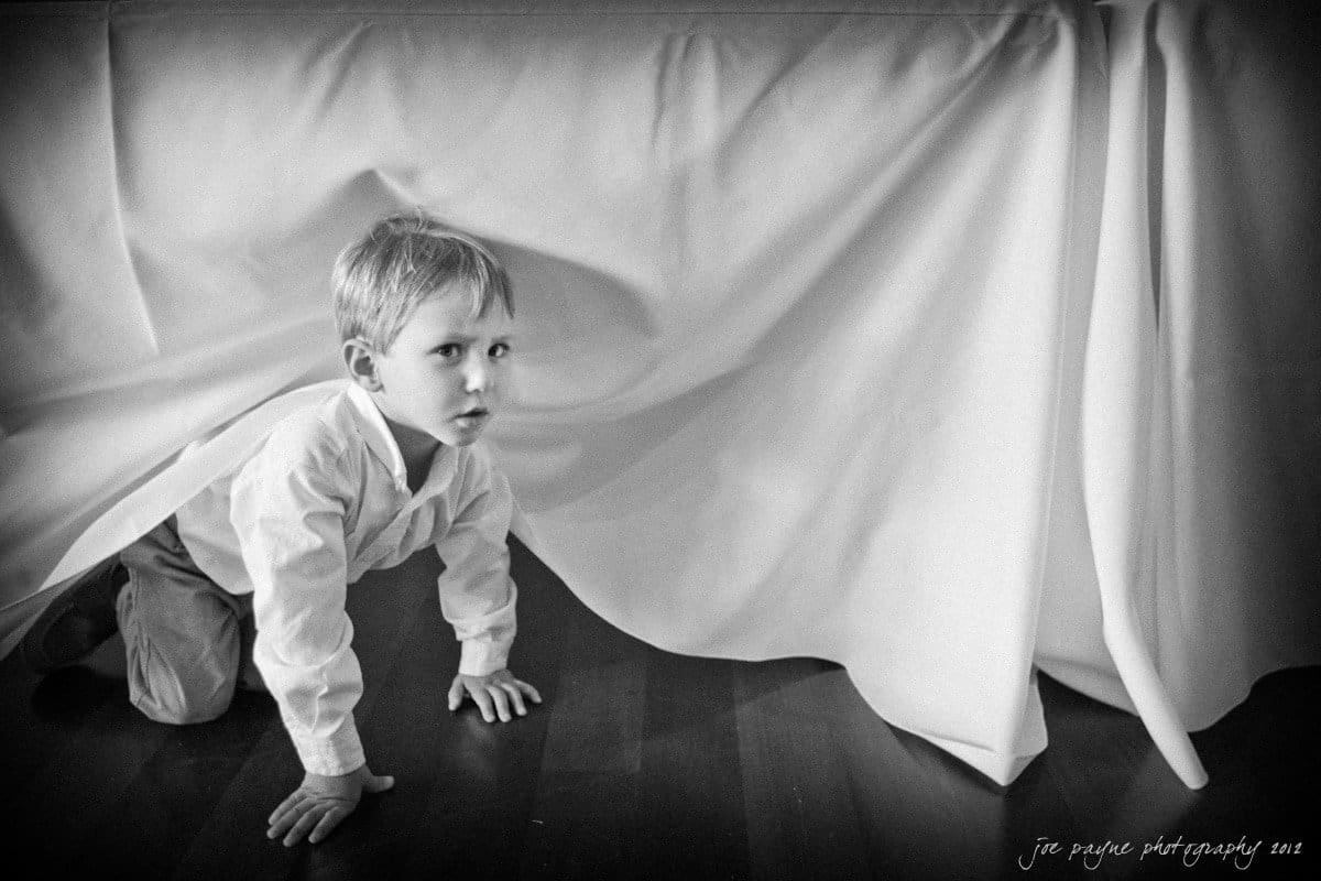 barn at valhalla, chapel hill wedding photography ~ allegra and chris A young child in a white shirt and pants crawls on a wooden floor, peeking from under a draped white tablecloth. The scene is in black and white, capturing a curious and playful moment.