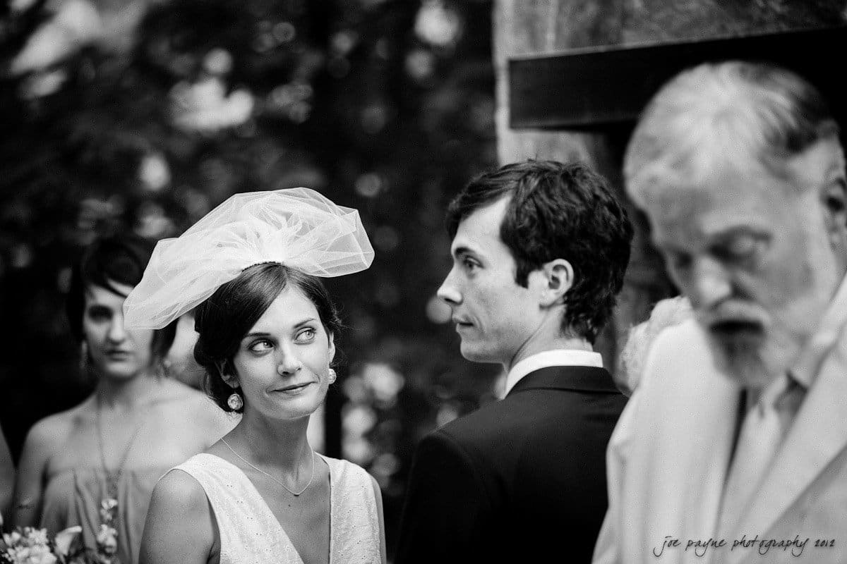 barn at valhalla, chapel hill wedding photography ~ allegra and chris A black and white photo of a wedding ceremony. A bride with a veil looks at the groom, who is facing a bearded man in a suit. A bridesmaid stands in the background. The scene is outdoors with trees visible in the background.