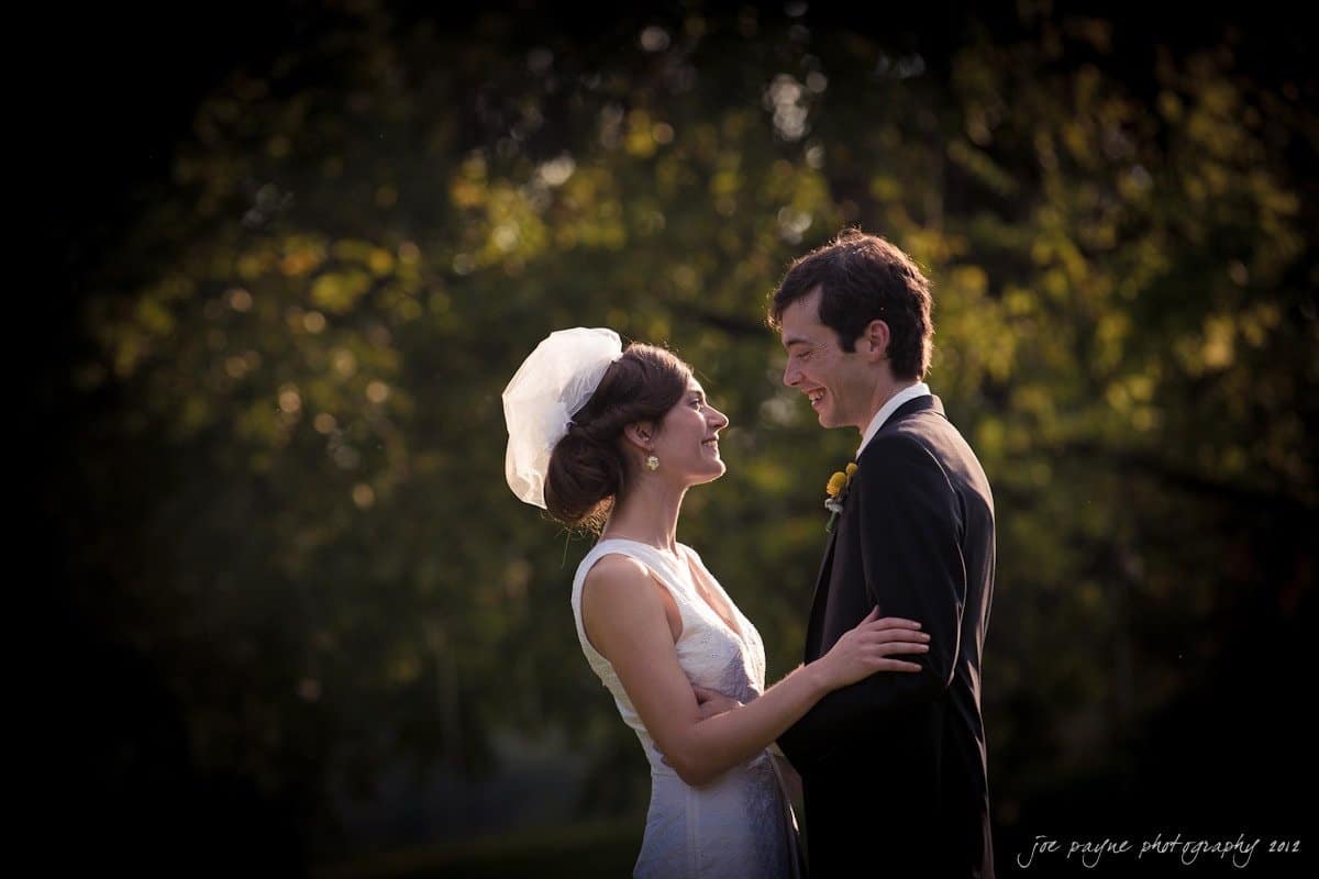 barn at valhalla, chapel hill wedding photography ~ allegra and chris A bride and groom share a joyful moment outdoors. The bride is in a white dress and veil, while the groom wears a dark suit with a boutonniere. They stand in front of a lush, green backdrop, smiling at each other warmly.