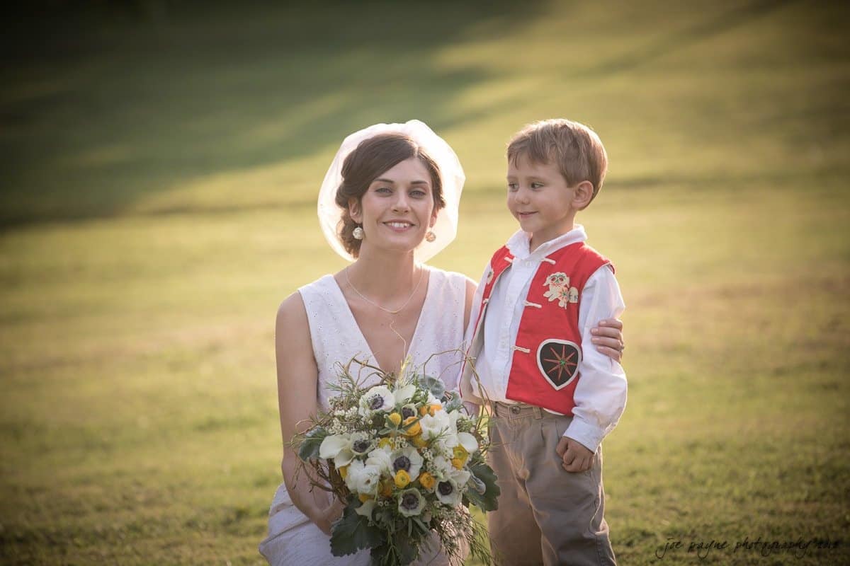 barn at valhalla, chapel hill wedding photography ~ allegra and chris A smiling woman in a white dress and veil kneels on grass next to a young boy in a red vest with embroidered flowers, white shirt, and beige pants. She holds a bouquet of yellow and white flowers. The background is a sunny, grassy area.