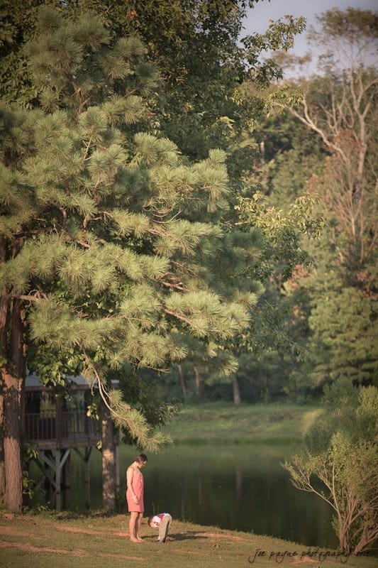 barn at valhalla, chapel hill wedding photography ~ allegra and chris A person wearing a red outfit stands on a grassy area near a large tree by a calm lake. A child, also in red, bends down next to them. The background includes a wooden structure and dense, green foliage.