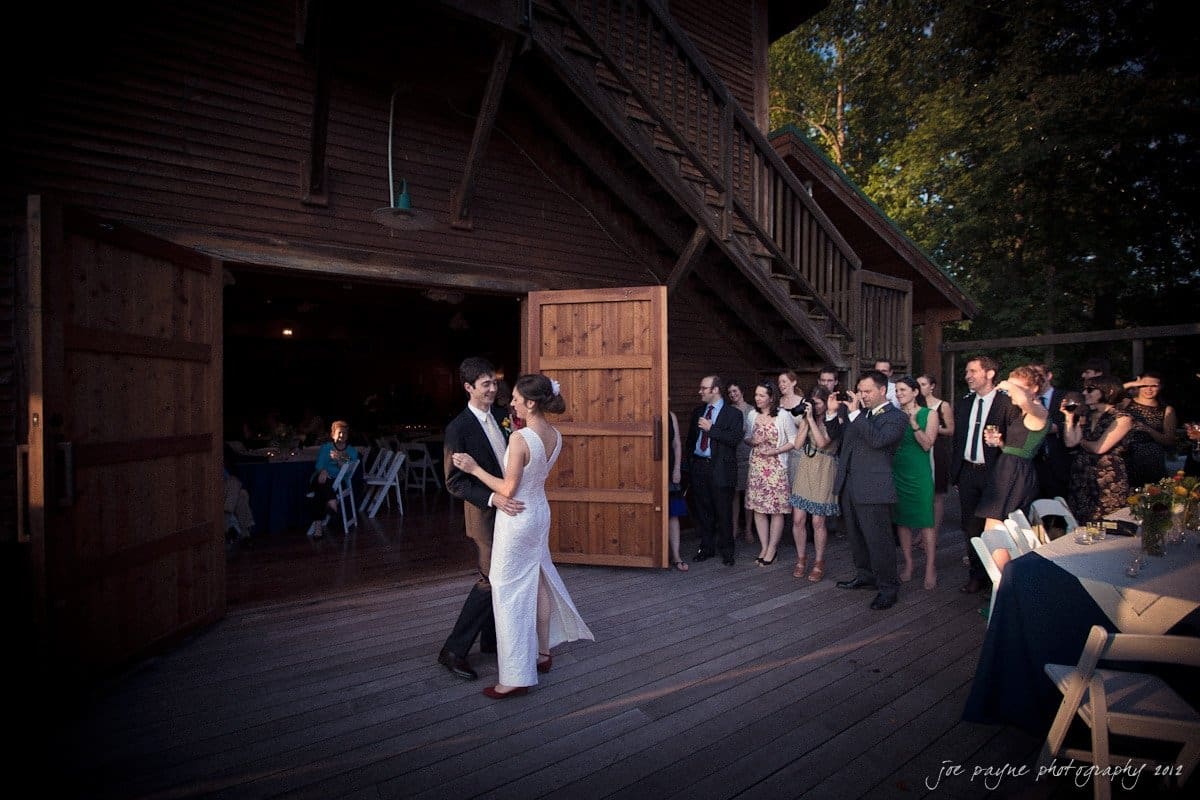 A couple dances on a wooden deck in front of an open barn. The bride wears a white dress, and the groom is in a black suit. Guests in formal attire stand nearby, watching and clapping. The setting is outdoors with trees visible in the background.