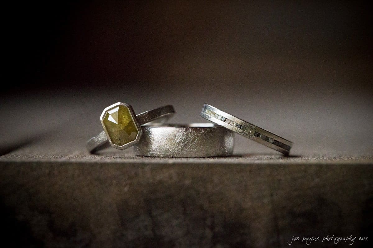 barn at valhalla, chapel hill wedding photography ~ allegra and chris A close-up of three silver rings on a textured wooden surface. One ring features a yellow gem, the center ring is plain, and the third ring has a subtle line design. The background is softly blurred, highlighting the rings.