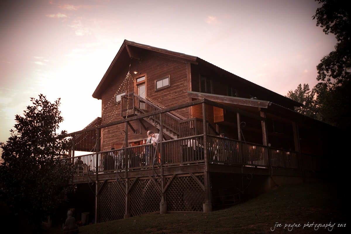 barn at valhalla, chapel hill wedding photography ~ allegra and chris A large wooden cabin with a wraparound porch is lit up at dusk. People stand on the porch, and soft lights are visible around the roof. The sky is painted with shades of orange and pink. Trees and grass surround the cabin.
