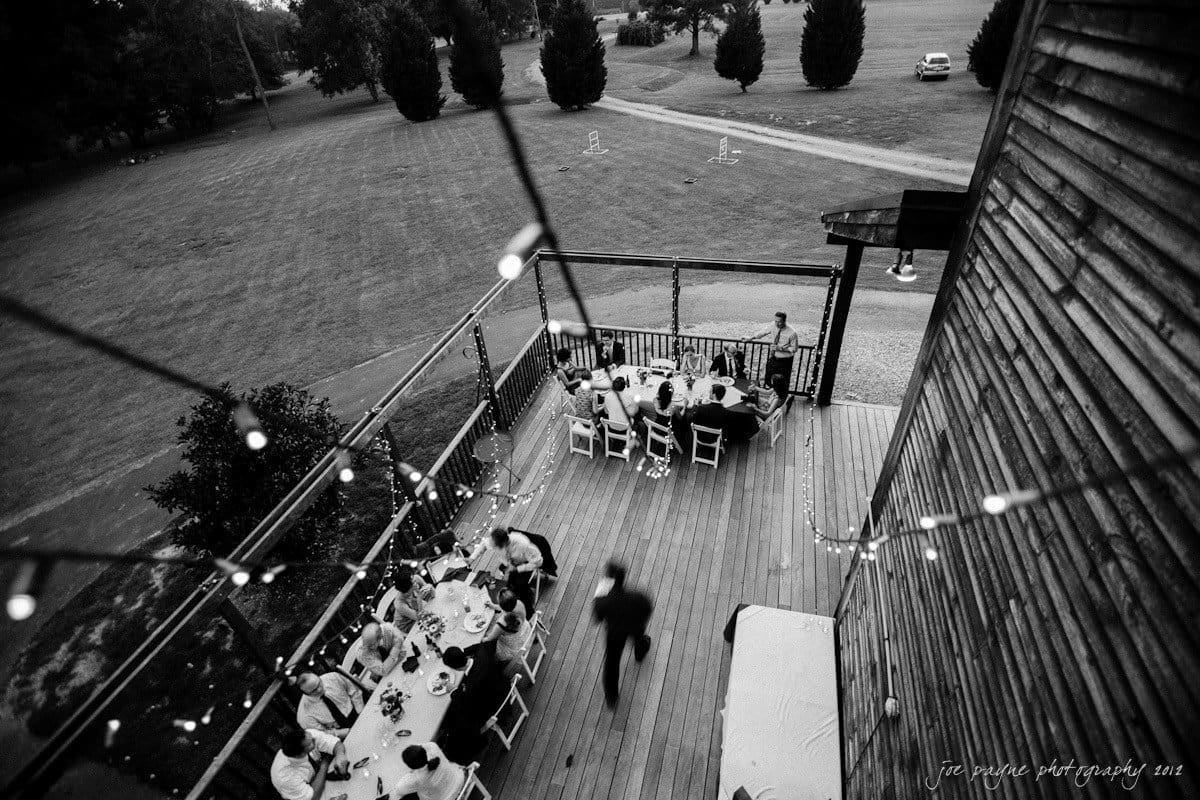 barn at valhalla, chapel hill wedding photography ~ allegra and chris A black and white photo of an outdoor gathering on a wooden deck. People are seated at tables, engaged in conversation. String lights are hung overhead, and a person is walking across the deck. A grassy field surrounds the area.