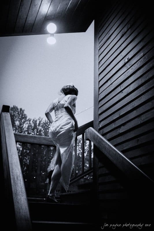 barn at valhalla, chapel hill wedding photography ~ allegra and chris A black-and-white photo of a person in a long dress ascending wooden stairs. The scene is dimly lit by a ceiling light above, with a backdrop of trees and a clear sky. The atmosphere is serene and reflective.
