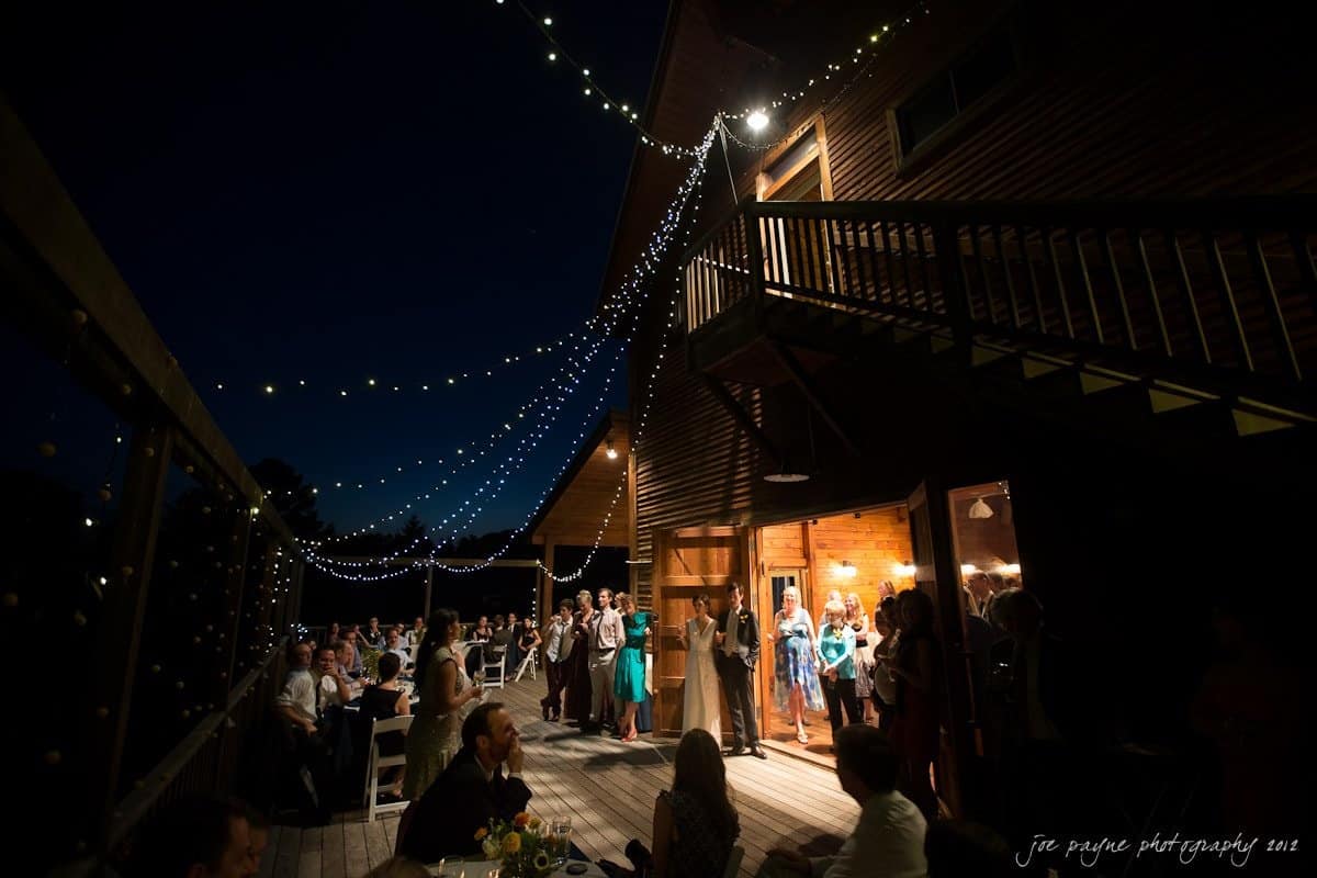 barn at valhalla, chapel hill wedding photography ~ allegra and chris People gathered at a nighttime outdoor event under string lights on a wooden deck. Some guests are seated at tables, while others stand near a warmly lit building. The sky is dark, and the mood is festive and relaxed.