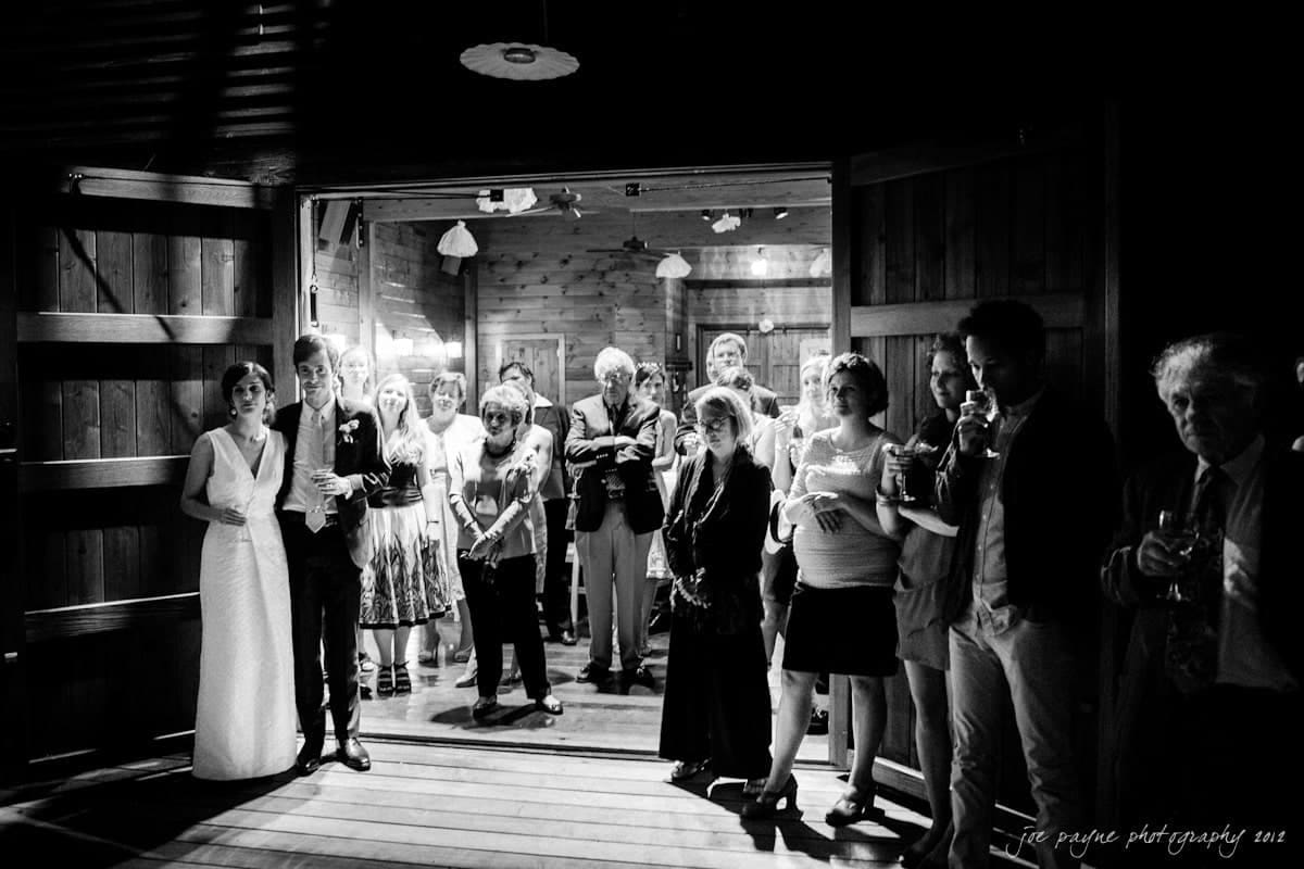 barn at valhalla, chapel hill wedding photography ~ allegra and chris A black and white photo of a wedding reception with a bride and groom standing to the left. Guests are gathered in the background, watching attentively. The room is dimly lit, with wooden decor and open doors.