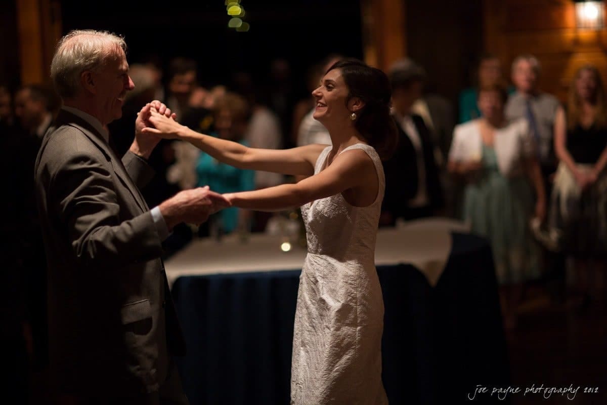 barn at valhalla, chapel hill wedding photography ~ allegra and chris A bride in a white dress dances with an older man in a suit at a wedding reception, surrounded by guests. The setting is warmly lit, creating a joyful and intimate atmosphere.