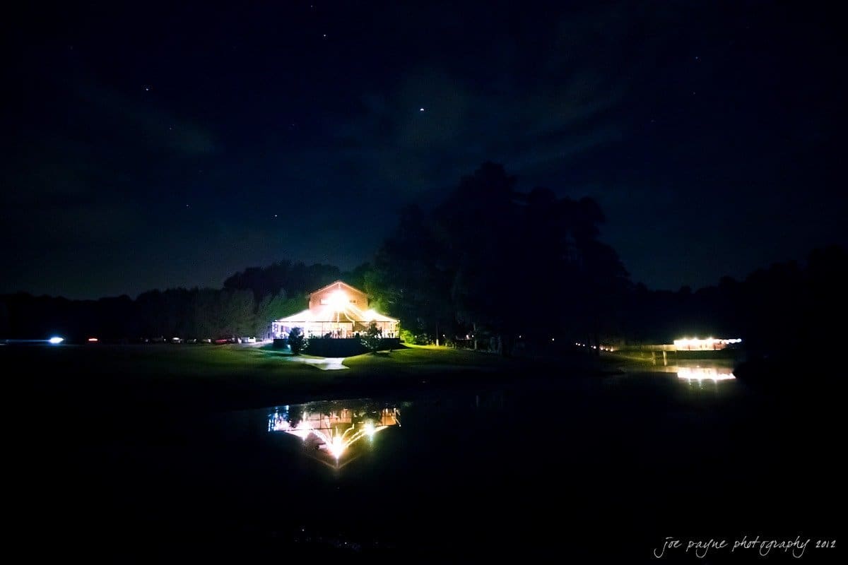 barn at valhalla, chapel hill wedding photography ~ allegra and chris A lit house stands by a lake, casting reflections on the water. The night sky is dotted with stars, and surrounding trees are silhouetted against the dimly lit background.