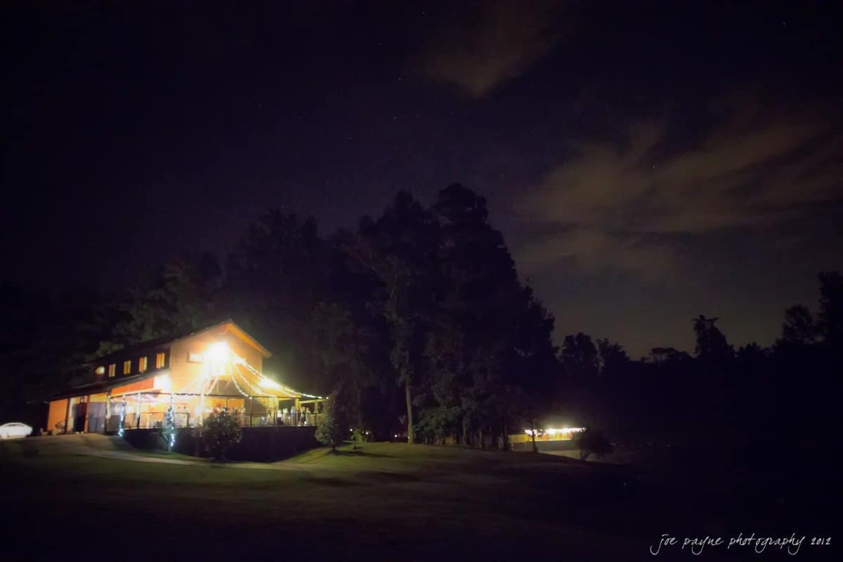 barn at valhalla, chapel hill wedding photography ~ allegra and chris A cozy, illuminated cabin is nestled among trees under a starry night sky. Soft lights shine from the windows, creating a warm glow. Shadows fill the surrounding grassy area, and a small light is visible in the distance.