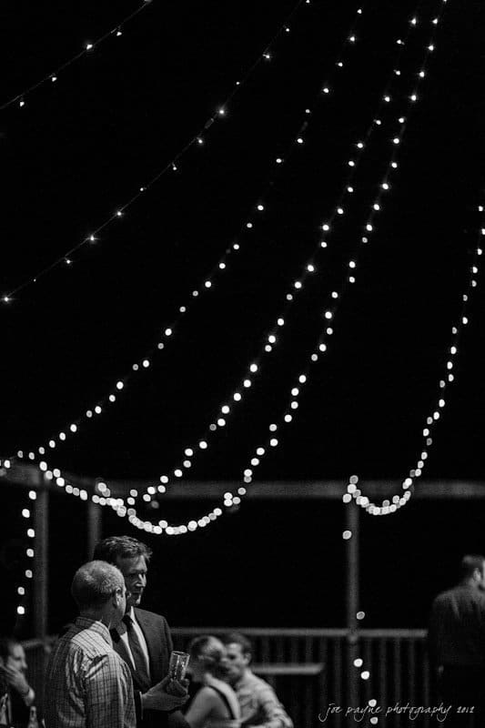 barn at valhalla, chapel hill wedding photography ~ allegra and chris A black and white image of people gathered at an event under strings of hanging lights. Two men are in the foreground, one in a suit, engaged in conversation. A group is seated in the background. The scene has an elegant, festive atmosphere.
